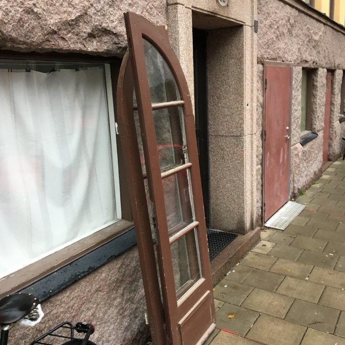 Brunmålad gammal dörr lutad mot en byggnad med stenfasad, målningen är sliten och visar dörrens åldringstecken.