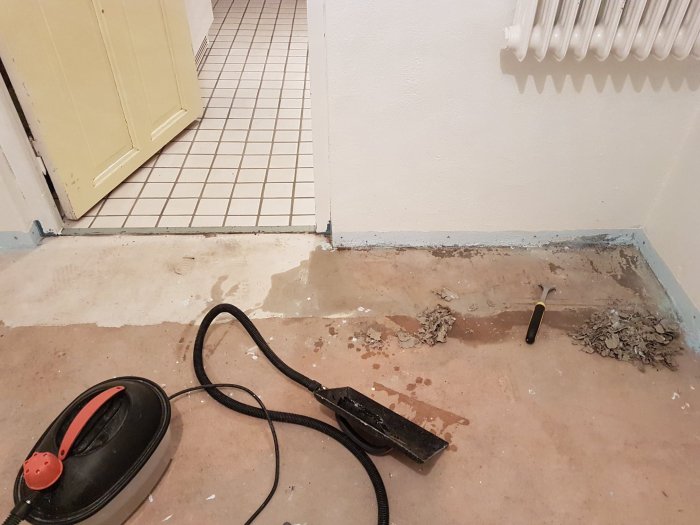 Källargolv med bortskrapade beläggningsrester och en dammsugarslang, skruvmejsel och skrapa.