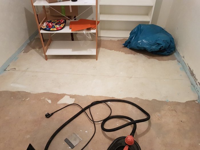 Källargolv med skrapade remsor av material, dammsugarslang och vit hylla i bakgrunden.