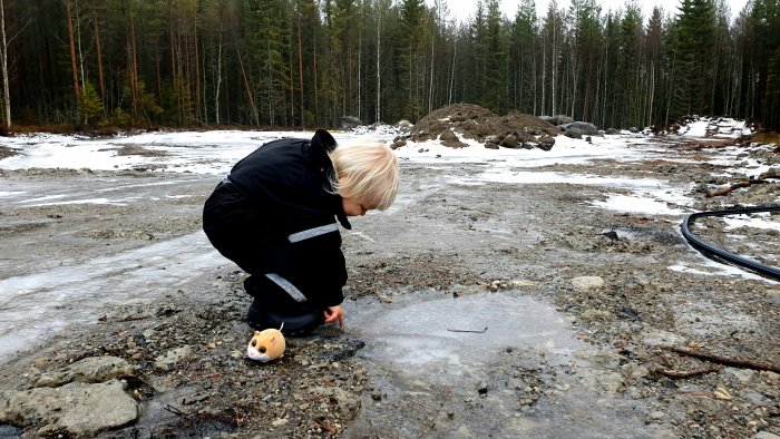 En person undersöker en isig tomtyta med träd i bakgrunden och en grushög, fotad från sydväst.