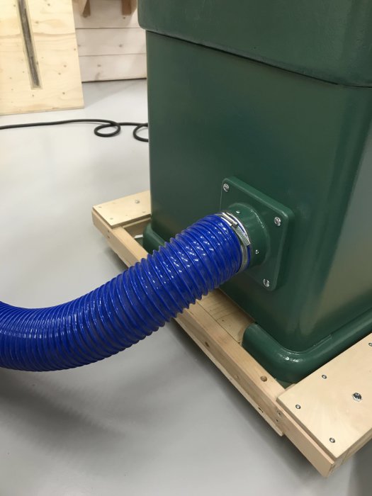 Uppgraderat spånutsug med 100mm blå slang ansluten till en grön maskin, monterad på en träplattform.