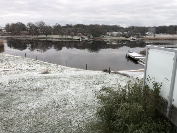 Vy med frostigt gräs, stilla sjö och bryggor mot bakgrund av träd och hus.