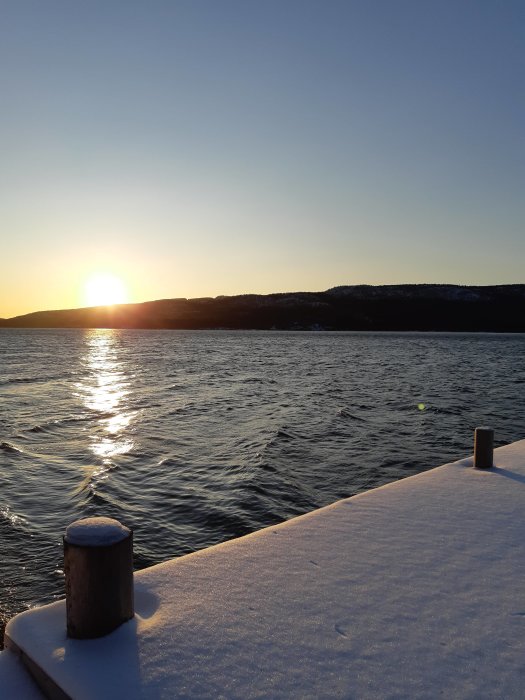 Blek vintersol nära horisonten, reflektion i vatten, snötäckt brygga med pollare.