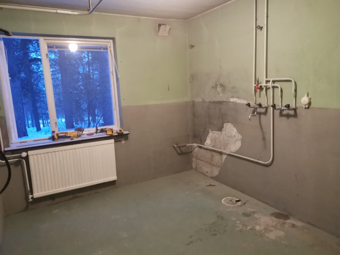 Rum under renovering med bortslipad väggfärg och exponerat fundament, rörkonstruktioner och ett fönster.