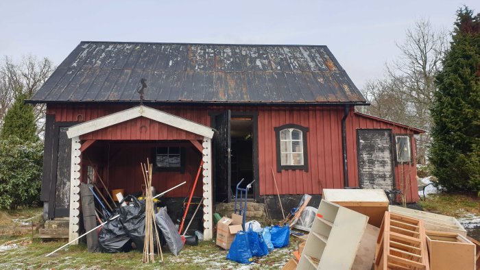 Rödmålat uthus med plåttak och diverse byggmaterial framför, samt en delvis borttagen utbyggnad.