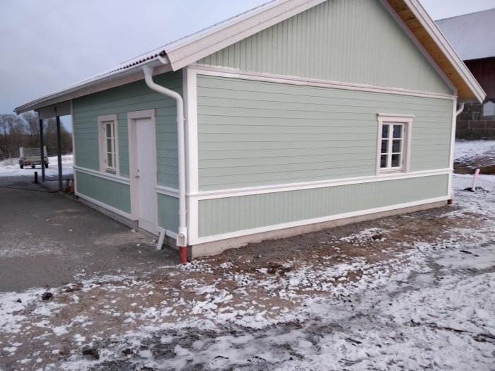 Nyinstallerade fönster och utvändigt foder på ett ljust grönt garage i dagsljus med lätt snötäcke på marken.