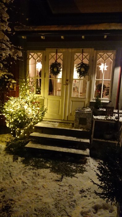 Entré med julpynt, upplyst buske och dörrkransar, snö på marken och adventsstjärnor i fönstren.