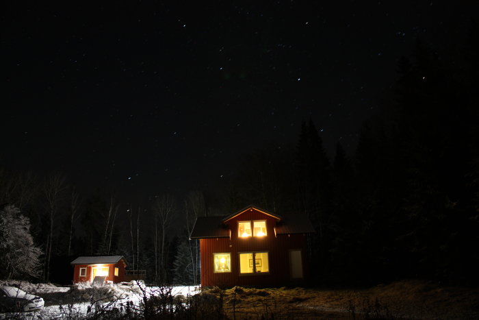 Nattbild av två belysta hus mot stjärnklar himmel med snötäckta träd i förgrunden.