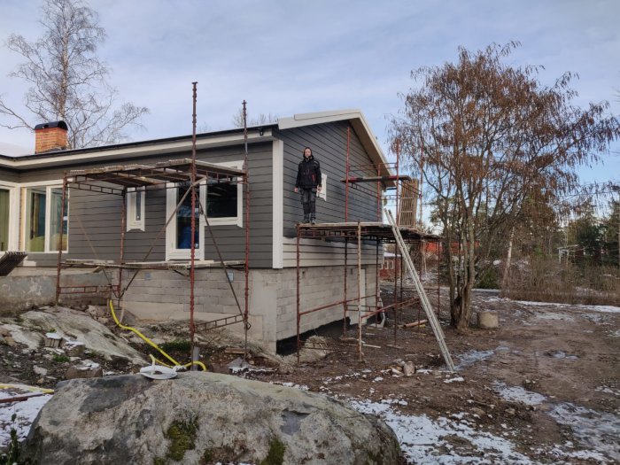 Ett hus under renovering med en person på ställningen, snöfläckar på marken och avklippt bladlös vegetation i bakgrunden.