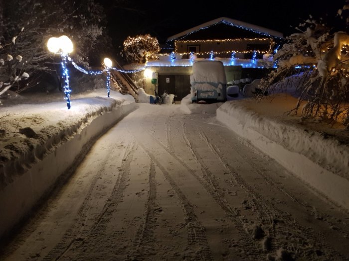 Snötäckt uppfart med julbelysning och ett täckt fordon, taget på natten.