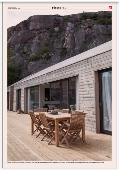 Huset med obehandlad träfasad och stora fönster mot en klippa, fört med ett träbord och stolar på terrassen.