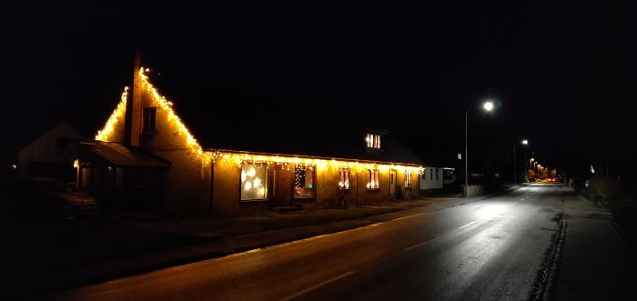 Ett hus dekorerat med julbelysning längs takkanten och fönster med julmotiv på natten.