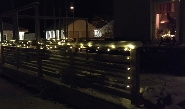 Ett altanräcke med en LED-slinga lindad runt, inbäddad i nysnö, och ett hus med adventsbelysning i bakgrunden.