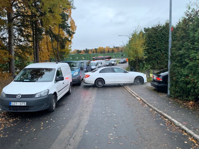 Bilar parkerade längs en smal gata, potentiellt blockerande utfart med träd och häck i bakgrunden.