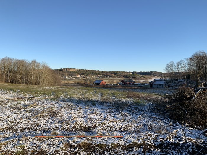 Landskapsvy över en dal med lätt snötäcke, hus i bakgrunden och soligt väder.