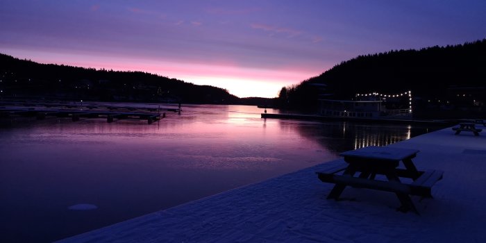 Morgongryning över en isbelagd sjö med bryggor och bänkbord under en livlig rosa och lila himmel.