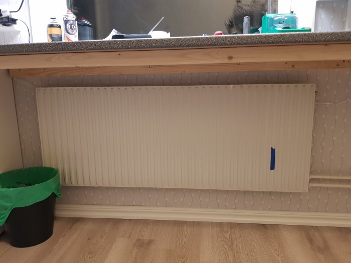 Radiator med blå tejp markerad 6-del varm, under hylla och bredvid sopkorg.