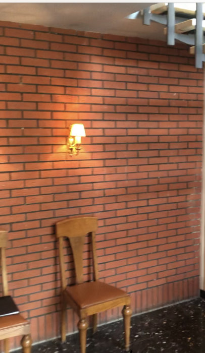 Entré med en tegelvägg i ljus nyans och en vägglampa, bredvid står en trästol.