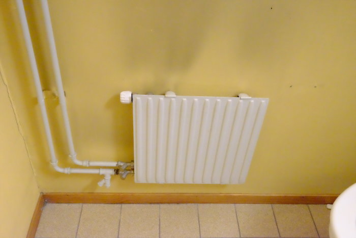 Väggmonterad vit radiator med synliga rör längs en gul vägg.