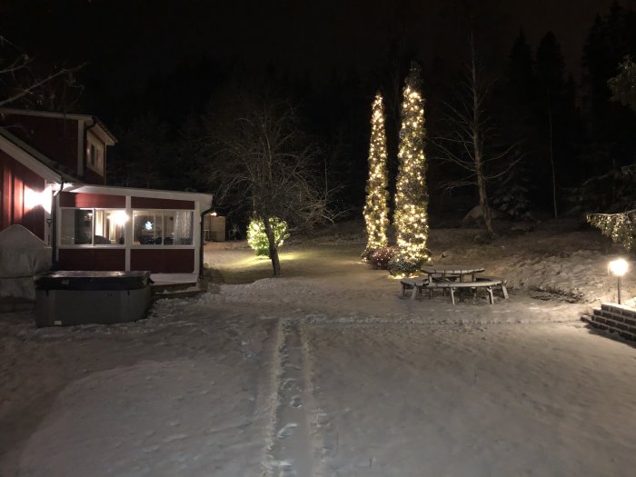 Vinterträdgård på natten med två upplysta Tujor täckta med lampor, snö, ett utebad och snötäckt picknickbord.