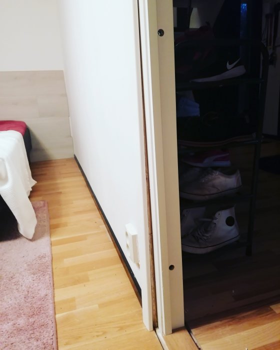 Sned dörrkarm intill en vit vägg och parkettgolv med en synlig glipa nedtill, nära ett skoställ.