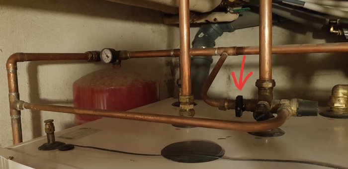 Kopparledningar med manometer, expansionskärl, och röd pil som pekar på en ventil för påfyllning i en källare.