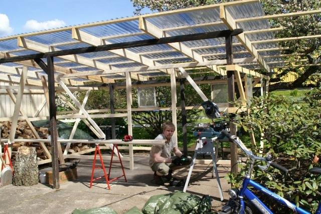 Under oppføring av et skur med gaflert tak, arbeider en person med verktøy på arbeidsplassen. Bouwelager og trær i bakgrunnen.
