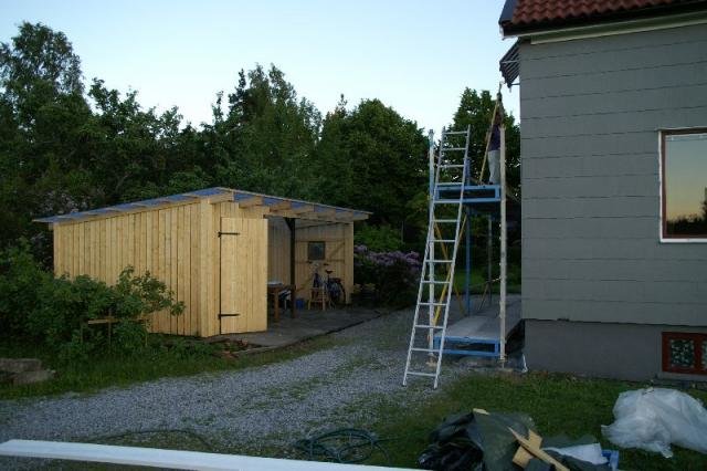 Et ferdigbygd skur ved siden av et hus med stige og arbeidsmaterialer i hagen.