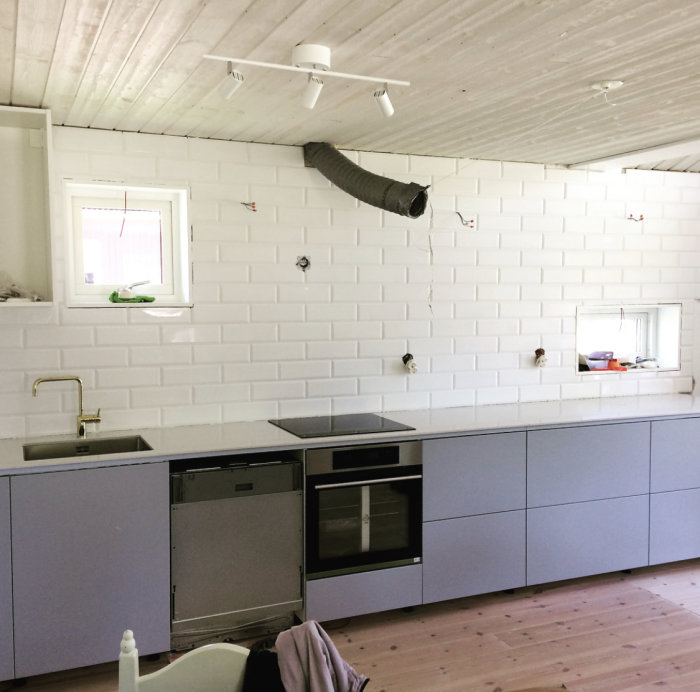 Renoverat kök halvfärdigt, nya skåp och kakel installerat, oavslutade eluttag och synlig ventilation.