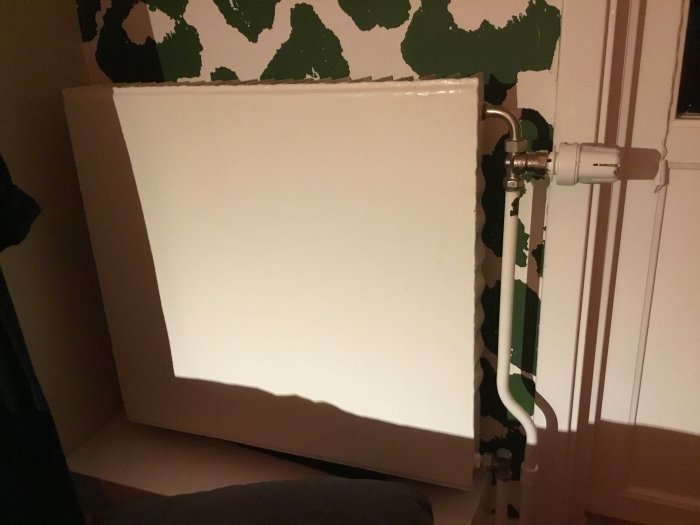 Radiator med termostatventil och anslutningar, placerad mot en vägg med avskalad tapet.