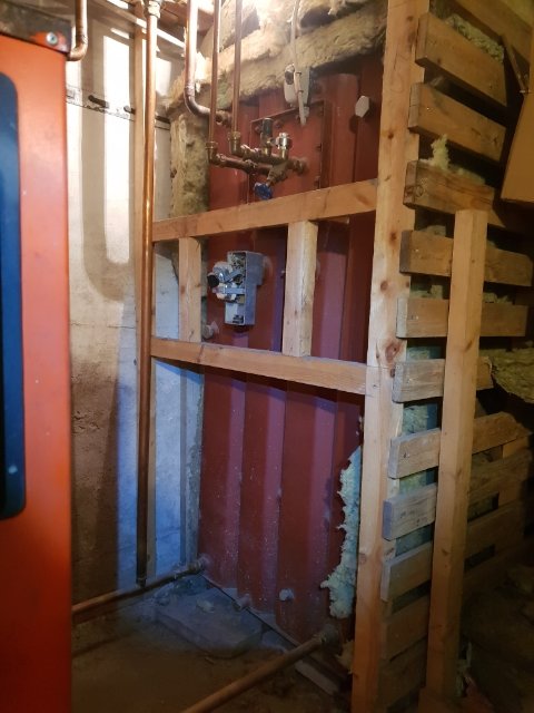 Värmeanläggning med synliga rör och ventiler i ett källarutrymme med oisolerade väggar och trästöttor.