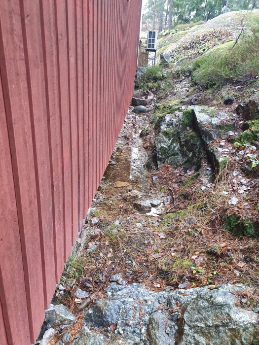 Vattenansamling längs krypgrund på sluttning med berg och faluröd husvägg.