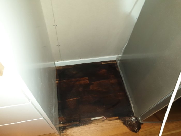 Vattenläcka och mögelskador under köksskåp med synligt mörkt trägolv och vit skåpdörr.