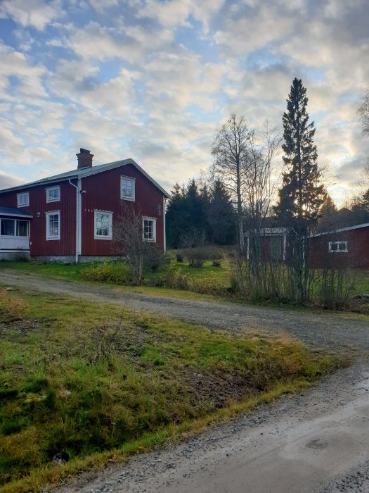 Röd trähusfasad med skorsten för vedspis, inramad av grönska och delvis molnig himmel.