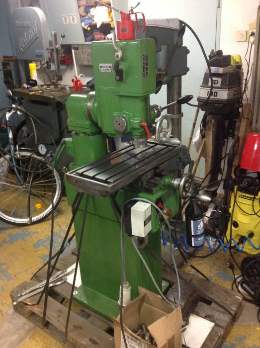 Grön fräs maskin med bord och olika handtag och rattar i en verkstadslokal med cykel och verktyg i bakgrunden.