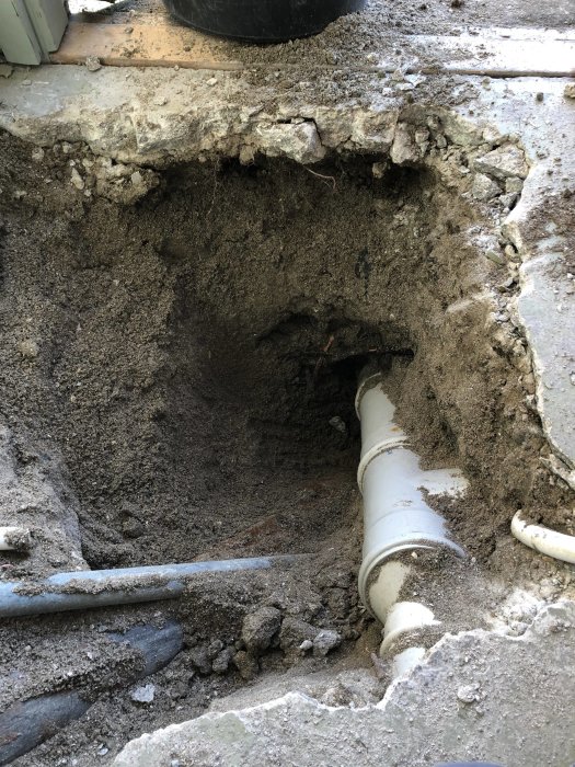 Grävd öppning som visar gammalt gjutjärnsrör anslutet till betongrör under källartröskel.