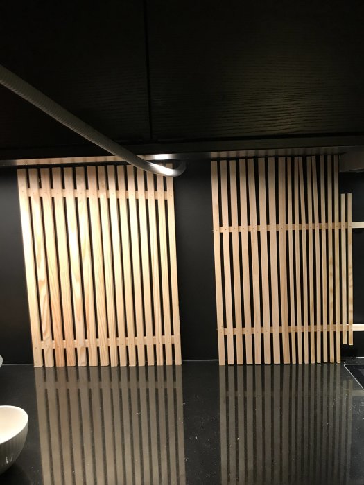 Träribbor av planhyvlat trä monterade på en svart vägg ovanför en diskbänk med reflektion på bänkskiva.