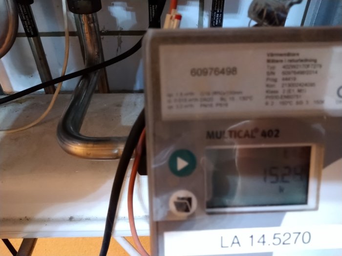 Närbild av en elektronisk värmeenergimätare vid ett värmesystem med rör och kablar i bakgrunden.