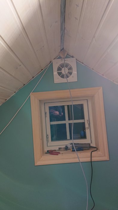 Ventilationsfläkt installerad på en lutande vitmålad takyta ovanför ett fönster i ett grönmålat rum.