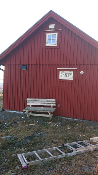 Röd stuga med ventilationsutrustning installerad, en vit bänk och nedfälld stege framför.