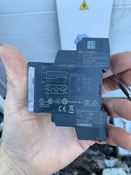 Hand håller ett Schneider Electric RE17RAMU tidrelä med tydligt märkta kopplingspunkter och ansiktschema.