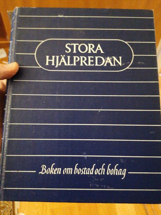 Hand som håller en blå gammal bok med titeln "STORA HJÄLPREDAN - Boken om bostad och bohag".