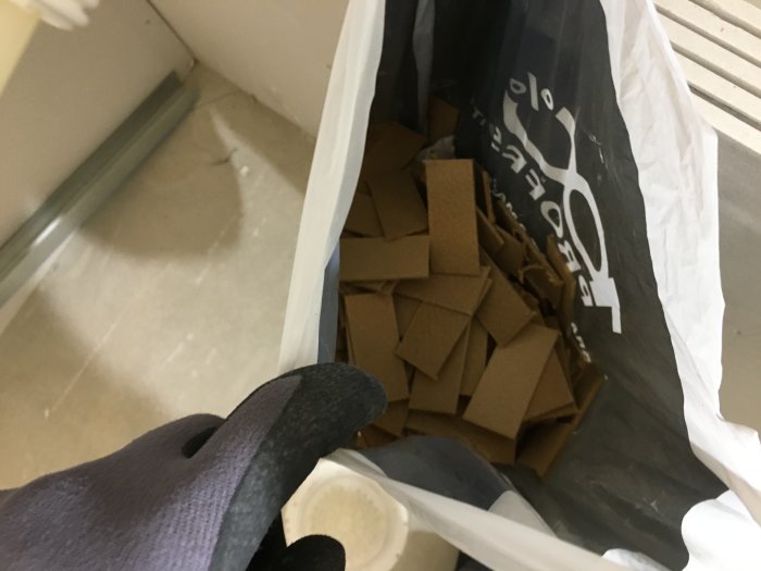 Vy över en påse fylld med masonitbitar på ett badrumsgolv, strategiskt placerade för att räta upp ett snett tak.