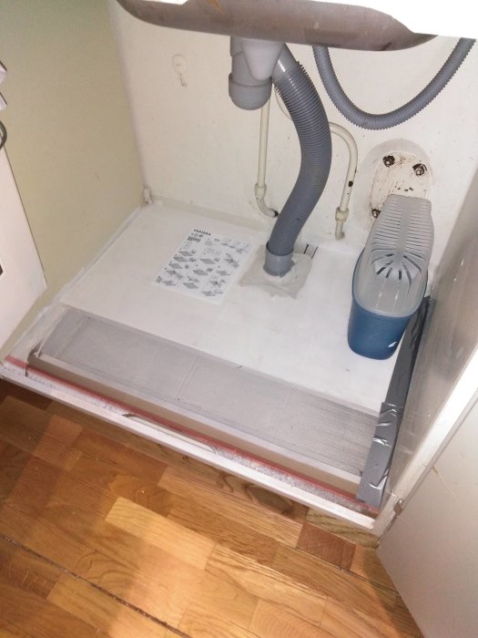 Nyinstallerad vit tvättmaskinsbotten med grå avloppsslang och blå lekagebehållare i skåp.