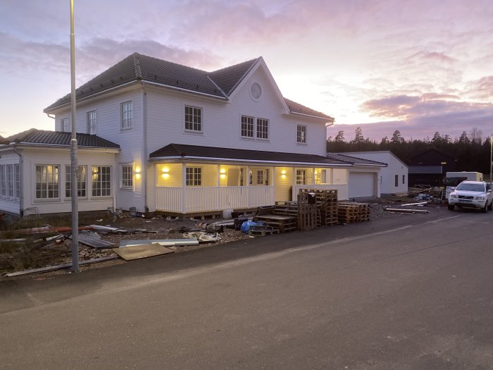 Renoveringsarbete utanför ett stort vitt hus vid skymning med byggmaterial och en bil parkerad.