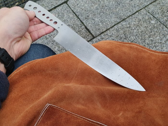 Hand som håller en obehandlad knivblad på ett brunt läderförkläde.