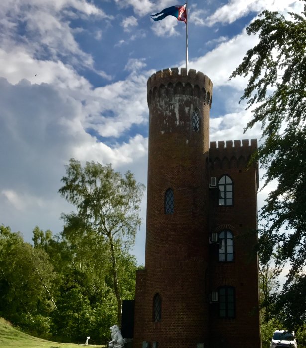 Miniatyr riddarborg med torn och tornflagga omgiven av grönska under blå himmel.