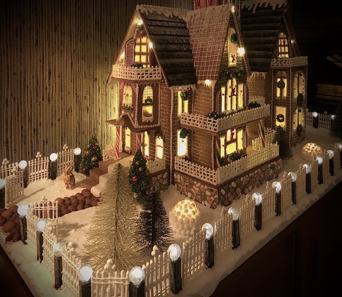 Ett detaljerat pepparkakshus med belyst staket, LED-belysning, tegelfasad, julgranar och snödekorationer.