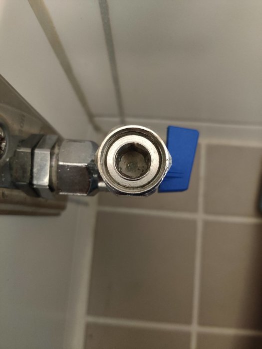 Närbild av en öppen kulventil med synlig bollmekanism mot en kakelbakgrund.