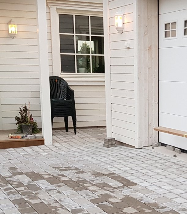 Stenlagd gång framför hus med vit panel, svart stol och vägglampor vid skymning.